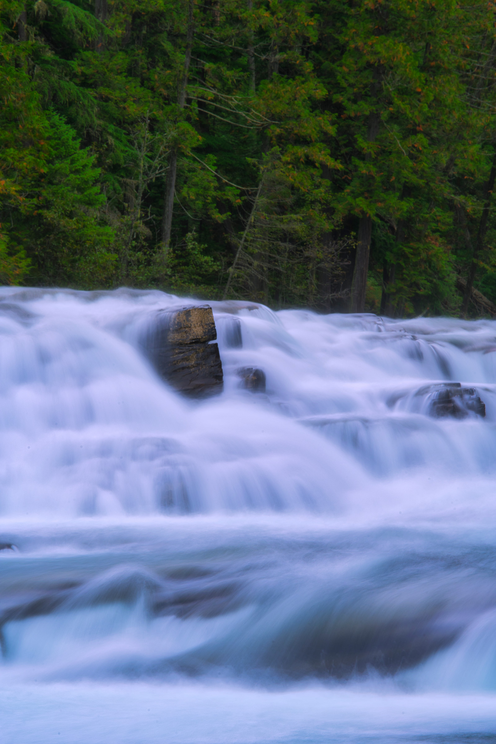 A powerful waterfall cascades over rocky ledges, with turbulent white water churning against dark stones in a lush green forest.