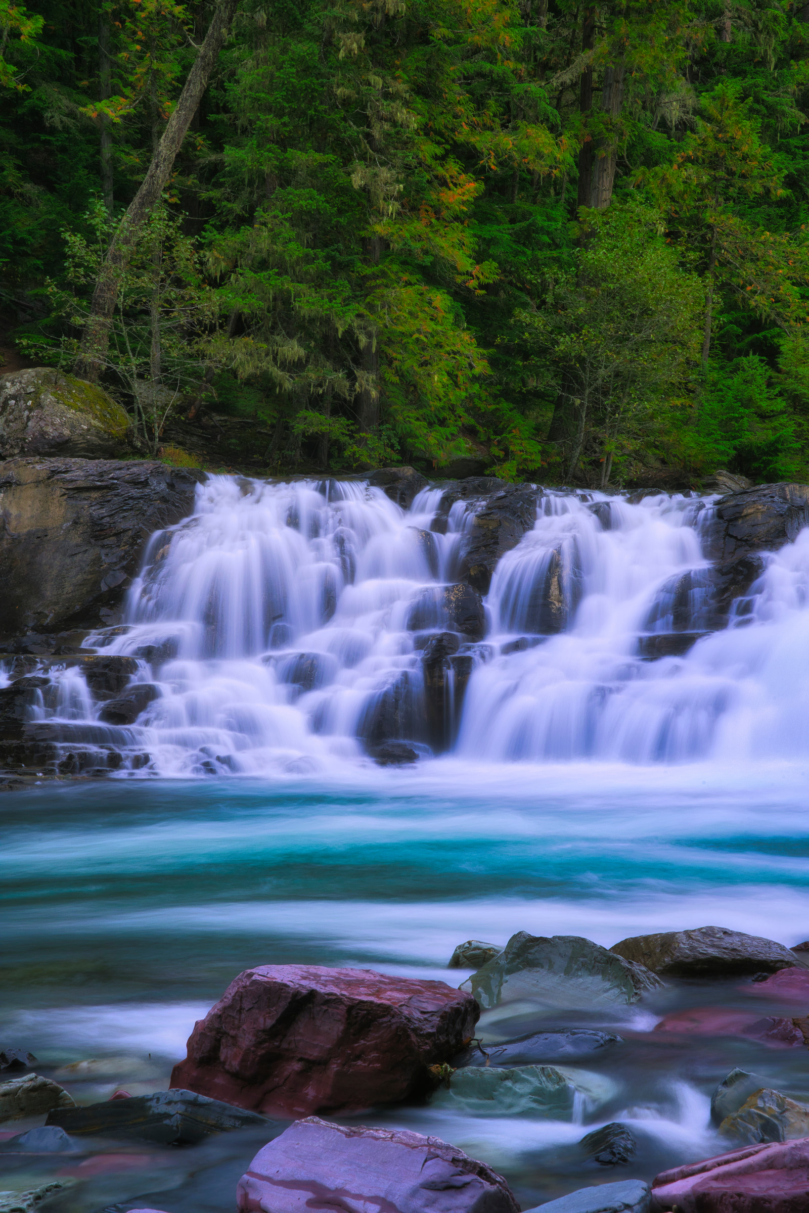 A waterfall cascades over rocky ledges into a turquoise river surrounded by dense forest.