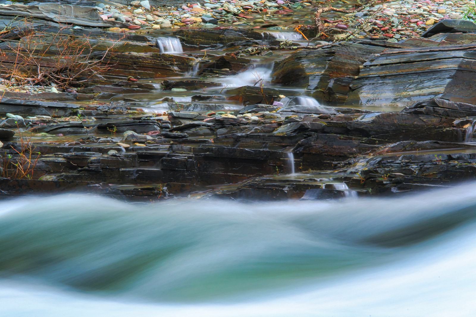 Rocky stream with small waterfalls cascading over layered stone steps, surrounded by scattered pebbles and sparse vegetation.