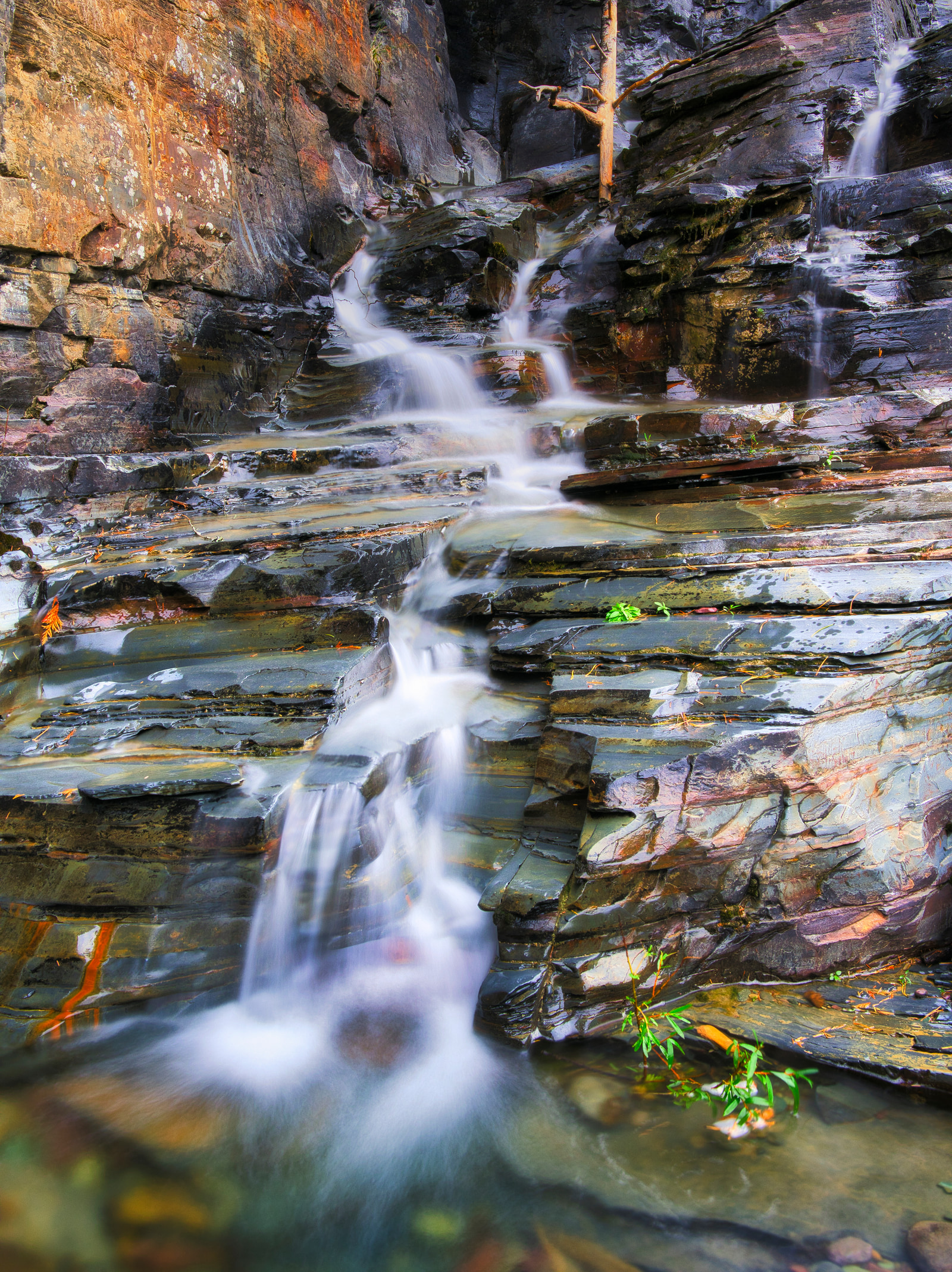 Steep, multi-tiered waterfall cascading over rugged, layered rock formations, with clear water flowing over mossy stones and small patches of greenery.