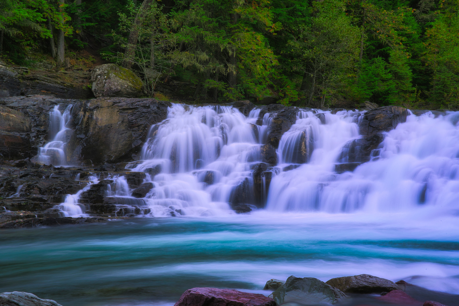 A multi-tiered waterfall cascades over dark rocks into a turquoise pool surrounded by lush green forest.