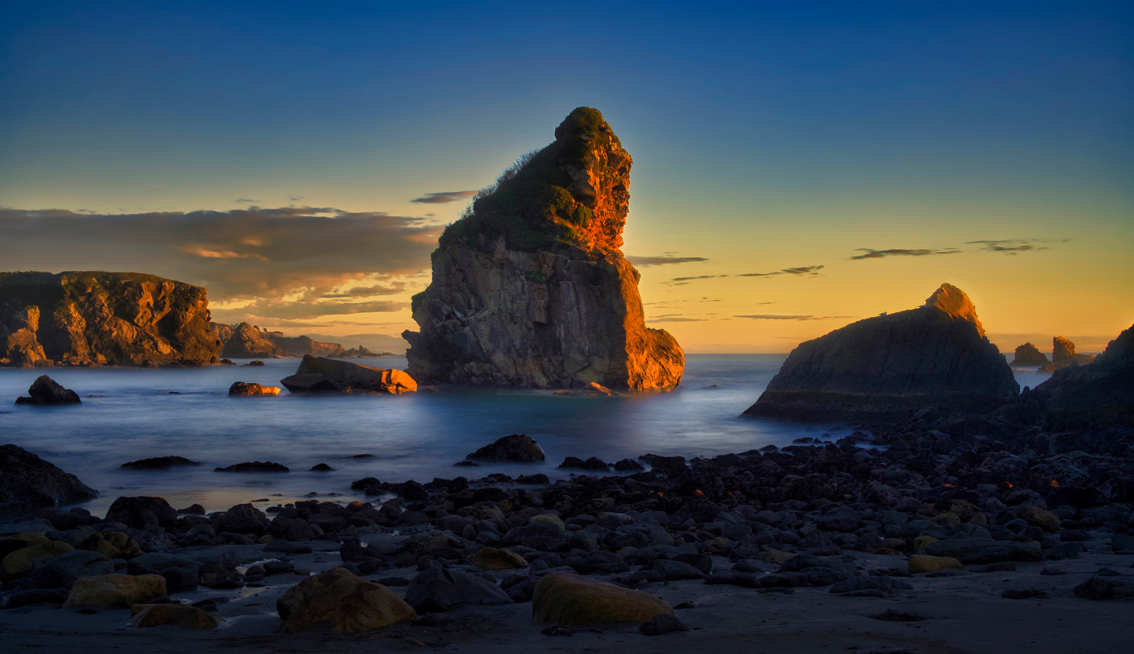 A rugged coastal rock formation stands prominently against a vibrant sunset sky, with calm turquoise water lapping against a rocky shore.
