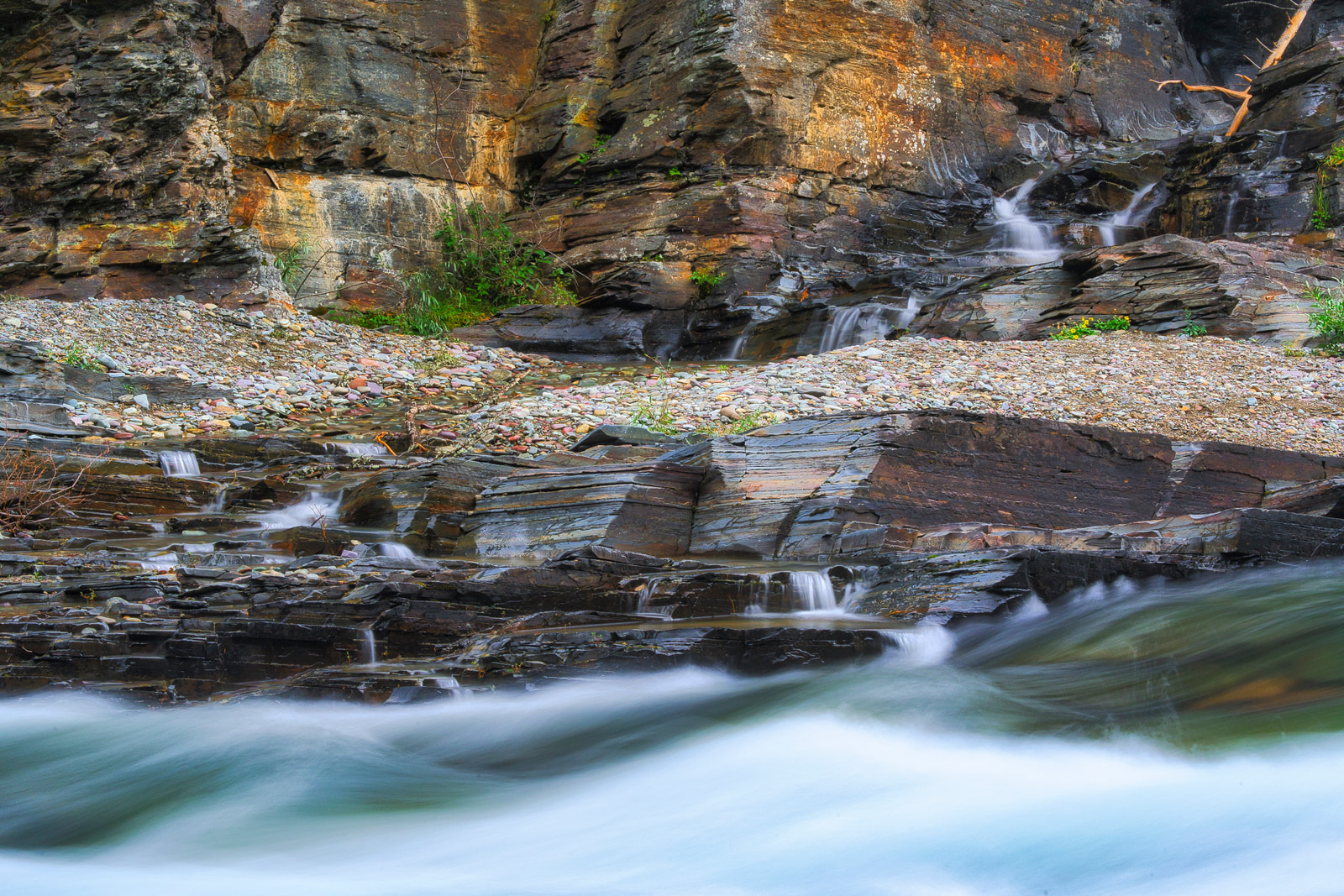 A rocky stream cascades over layered stone ledges, with small waterfalls and clear water flowing over mossy rocks.