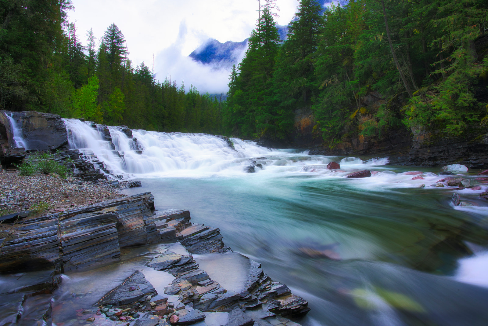 A waterfall cascades over rocky ledges into a turquoise river, surrounded by dense evergreen forest and misty mountains.