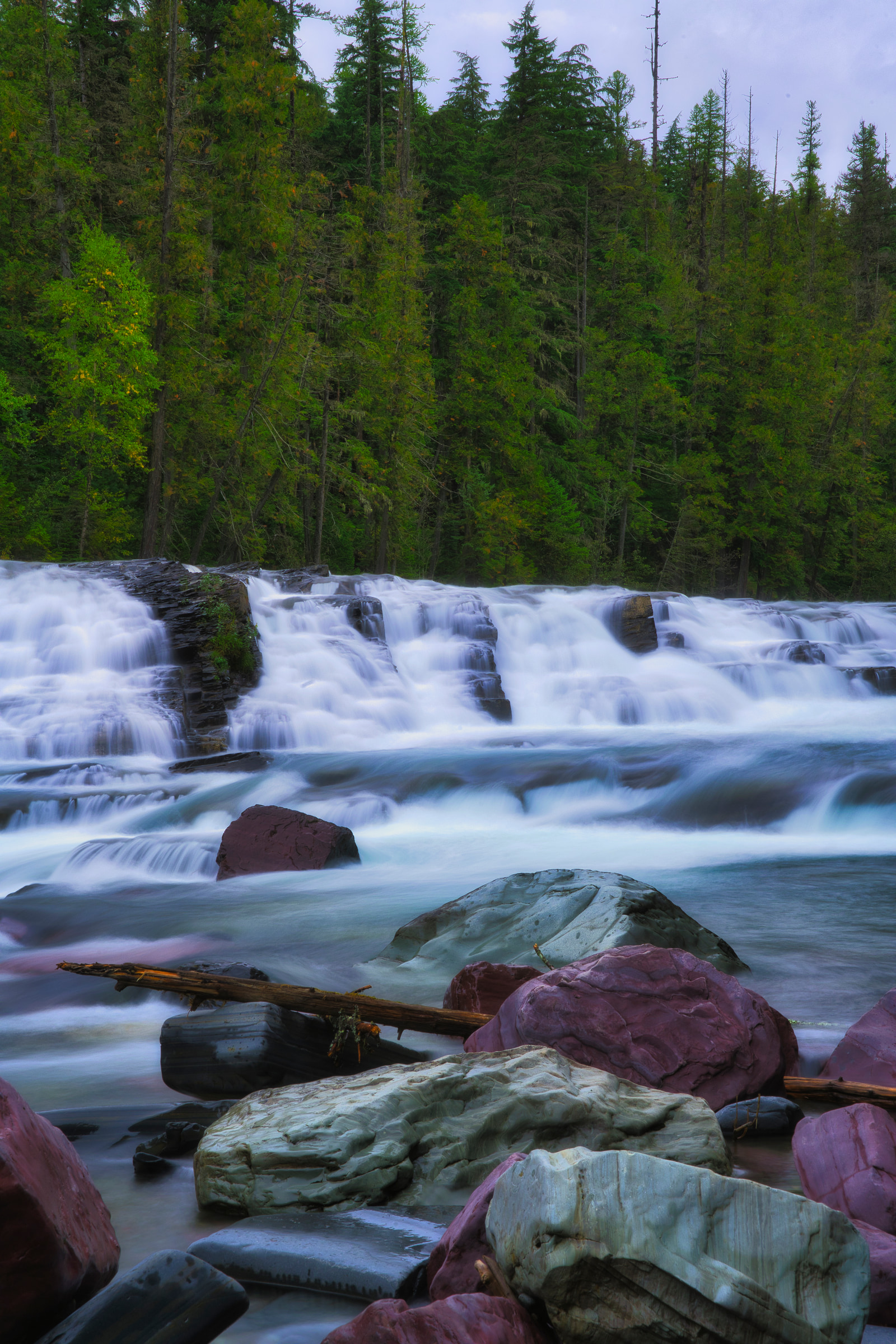A wide waterfall cascades over rocky ledges, with turbulent white water flowing into a pool below, surrounded by dense green forest.