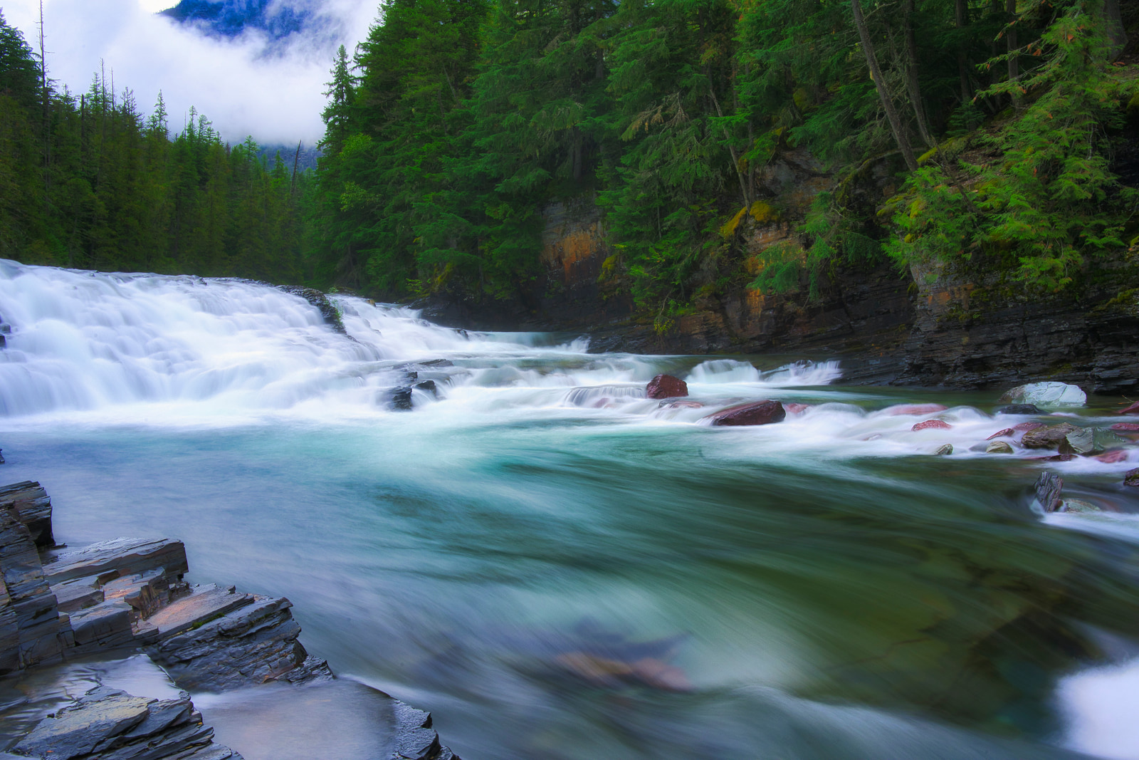 A waterfall cascades over rocky ledges into a clear river surrounded by dense evergreen forest under a misty sky.