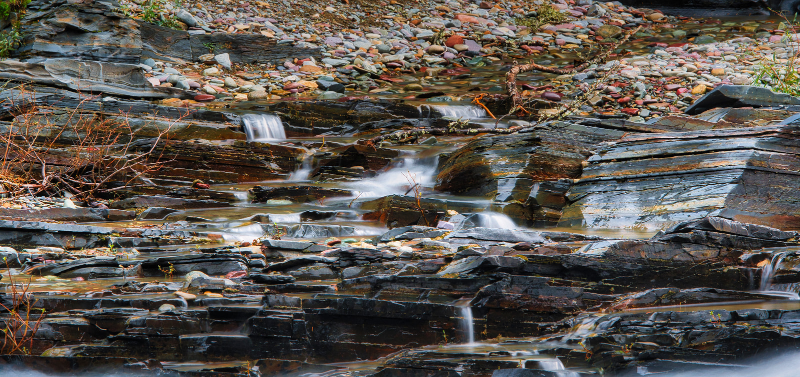 Rocky stream with small waterfalls cascading over layered slate, surrounded by scattered pebbles and sparse vegetation.