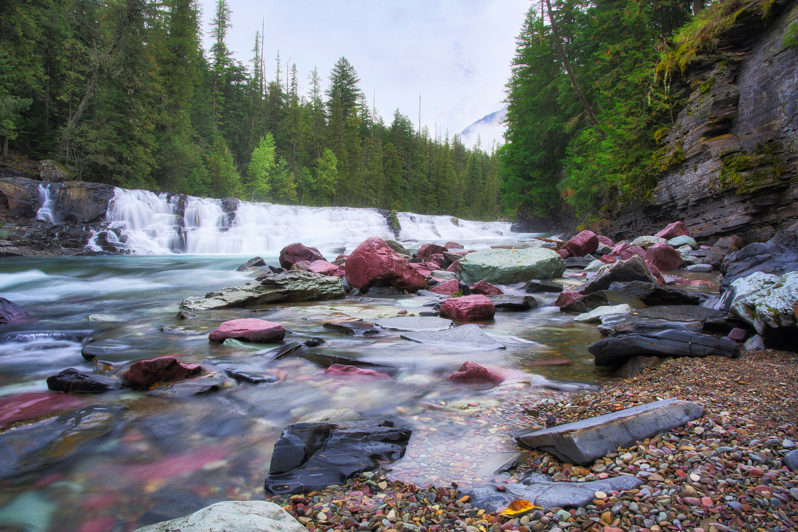 A waterfall cascades over rocky ledges into a clear river, surrounded by dense forest and scattered boulders.