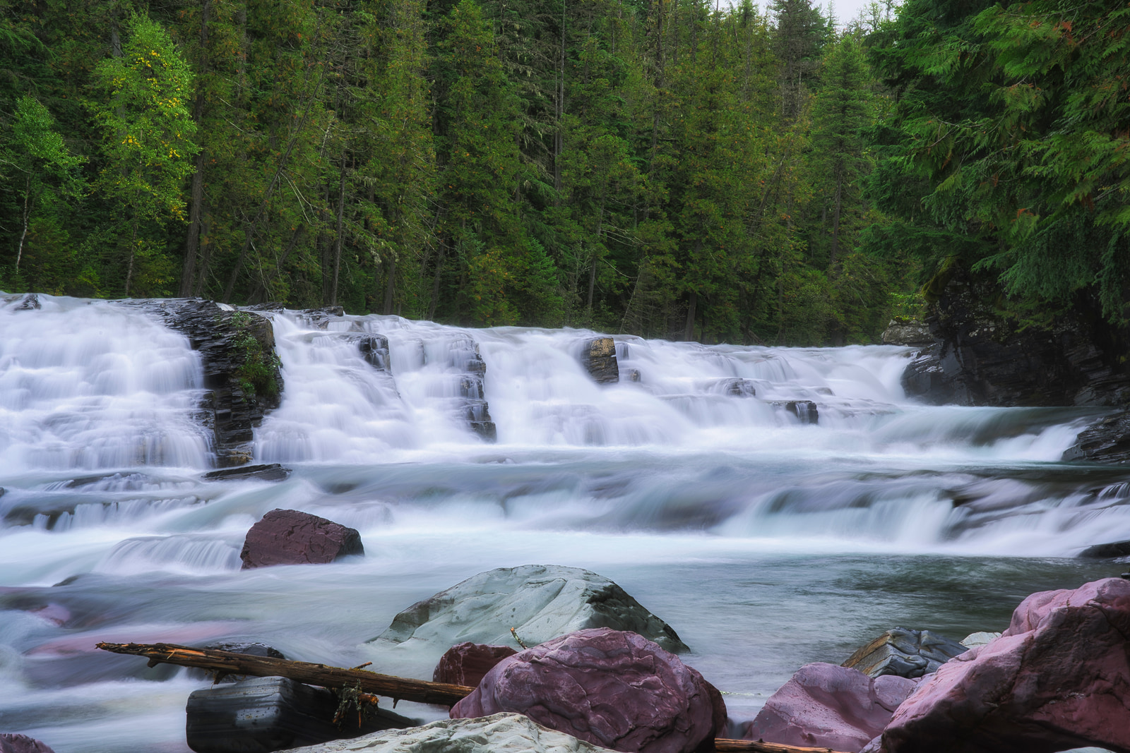 A wide waterfall cascades over rocky ledges into a fast-flowing river, surrounded by dense green forest.