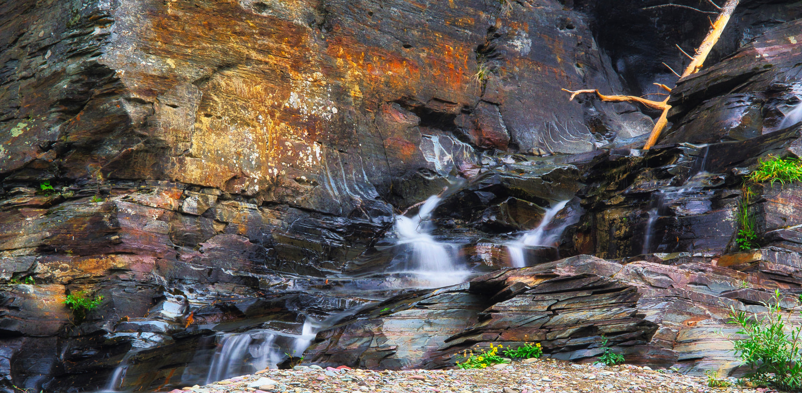 Rocky cliff with small waterfalls cascading down its face, surrounded by patches of green vegetation.