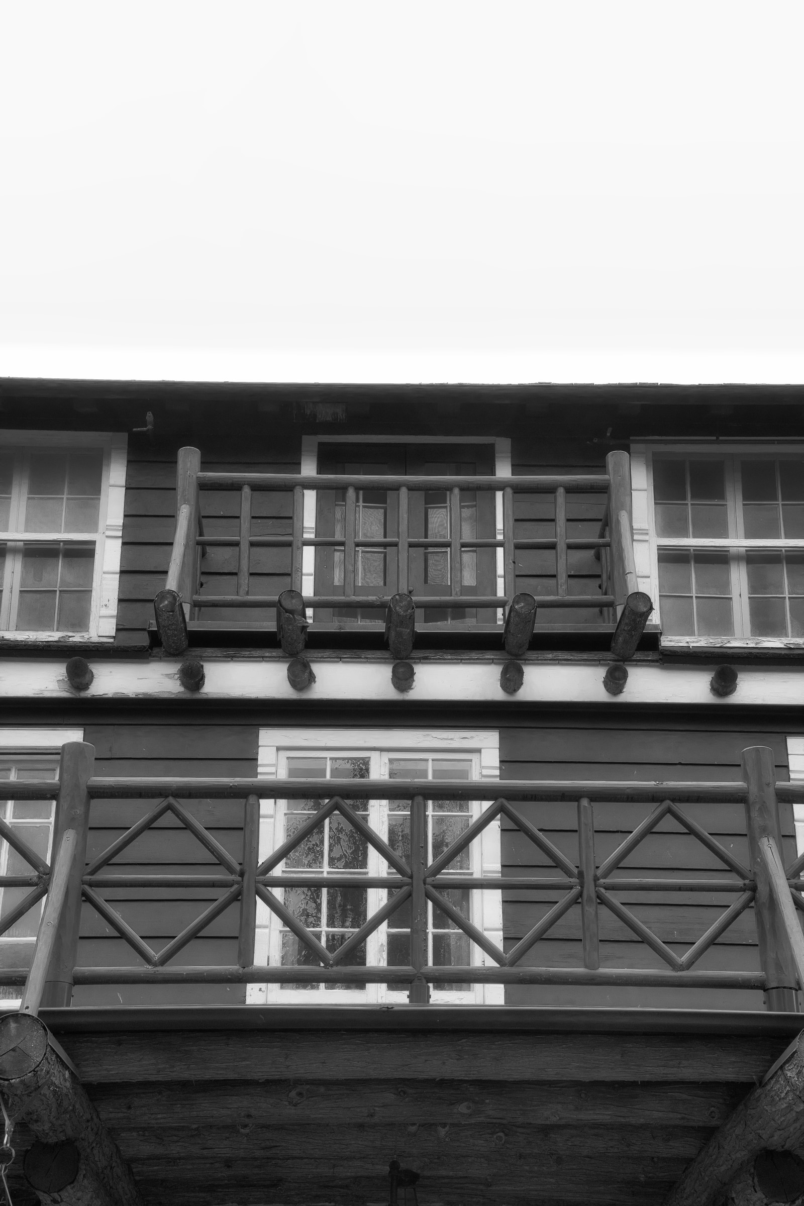 A black-and-white photograph shows a wooden balcony with a decorative railing and multiple windows on the building behind it.