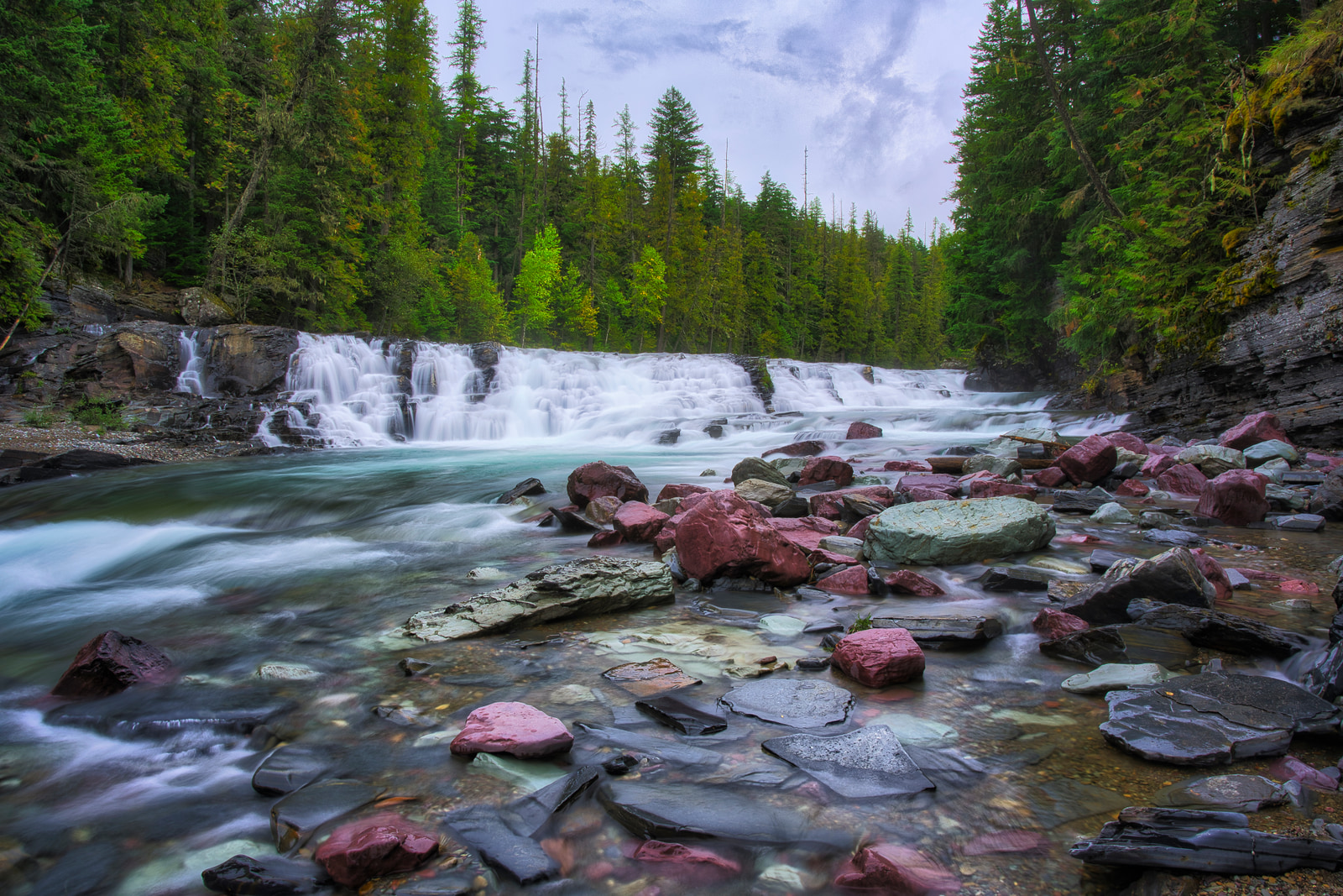 A waterfall cascades over rocky ledges into a clear river lined with colorful stones and surrounded by dense evergreen forest.