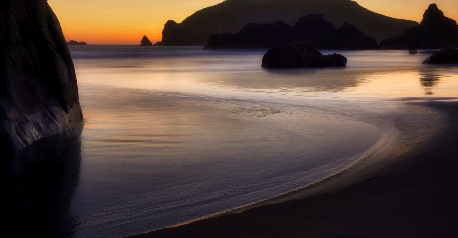 A tranquil beach at sunset with calm water reflecting the warm orange and pink hues of the sky, rocky outcrops silhouetted against the distant horizon.