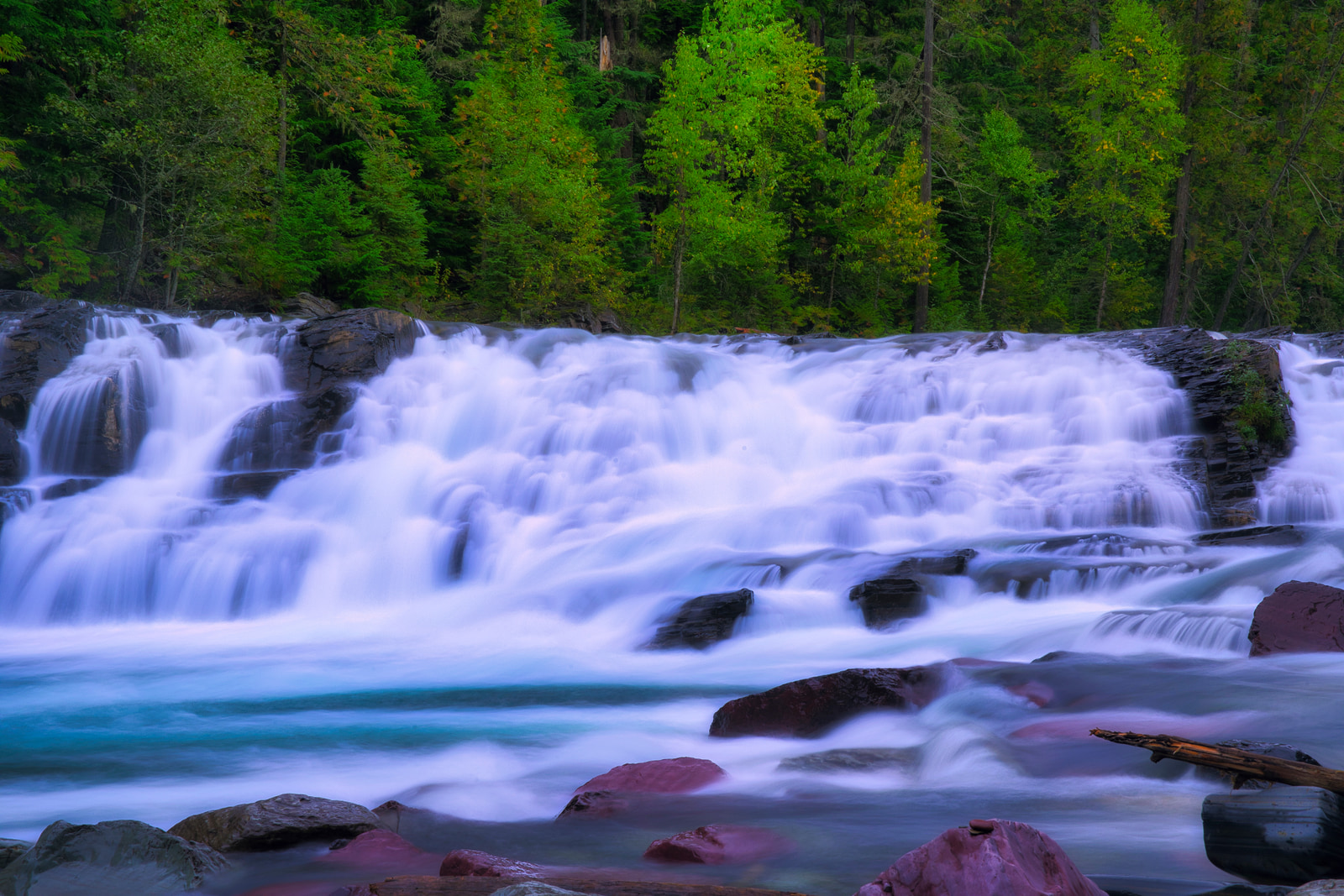 A wide waterfall cascades over rocky ledges into a clear pool below, surrounded by dense green forest.