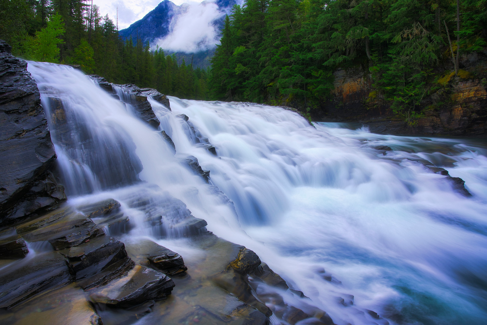 A powerful waterfall cascades over rugged rocks, its white water churning against dark stone in a lush, forested valley.