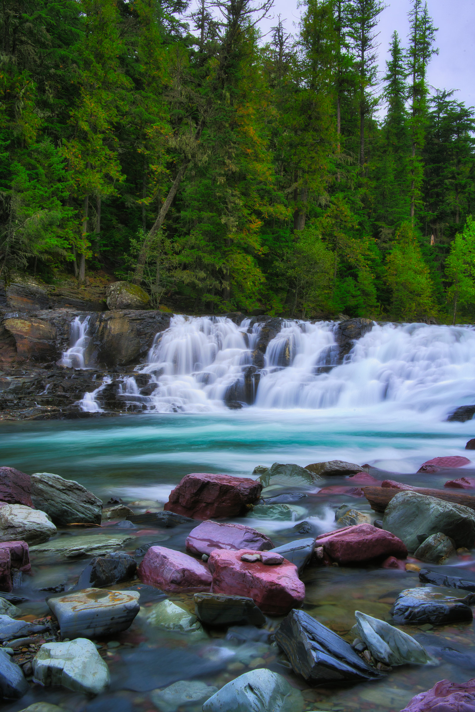 A wide waterfall cascades over rocky ledges into a turquoise river, surrounded by dense evergreen forest and scattered boulders.