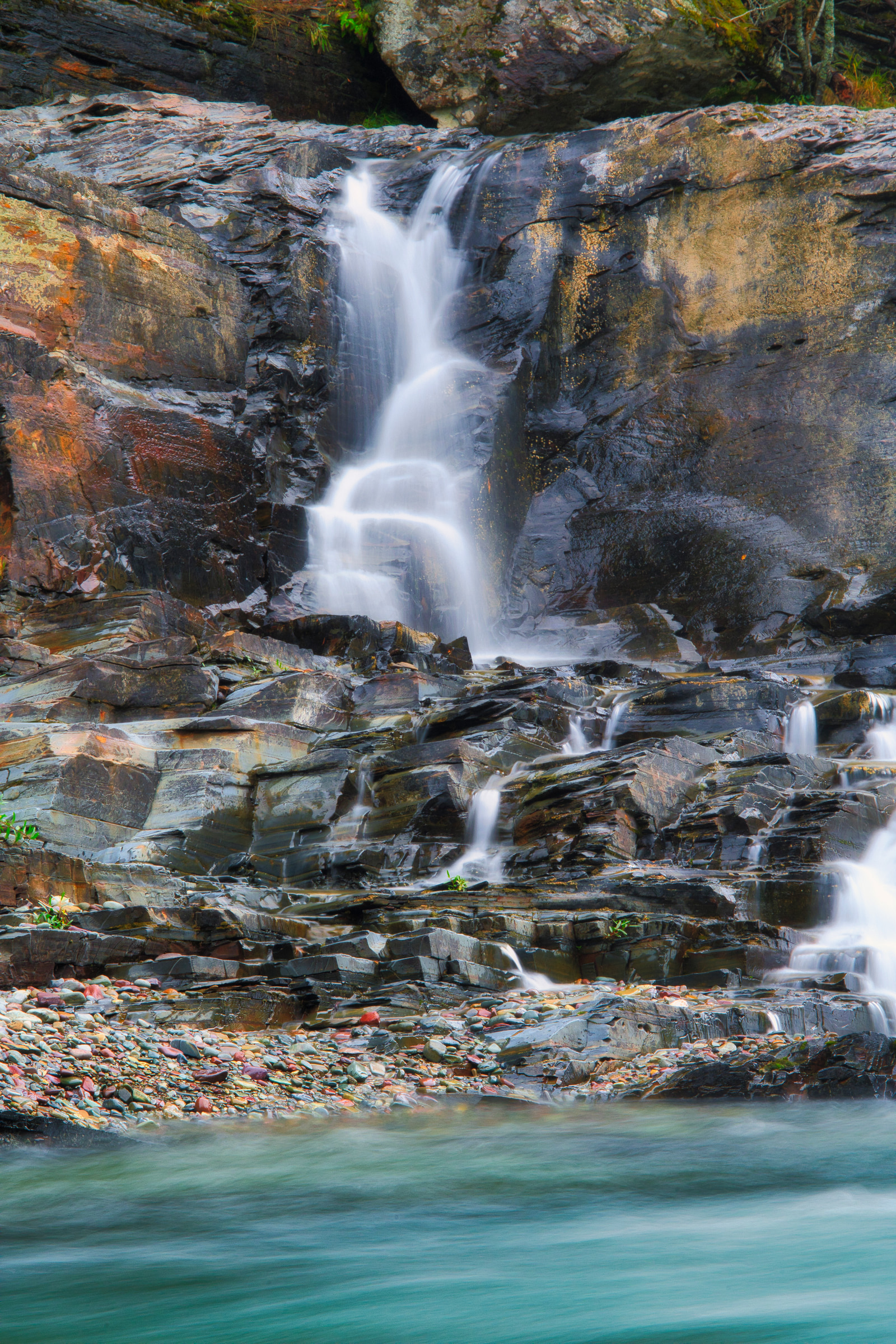 A multi-tiered waterfall cascades over rugged, layered rock formations into a clear turquoise pool below.